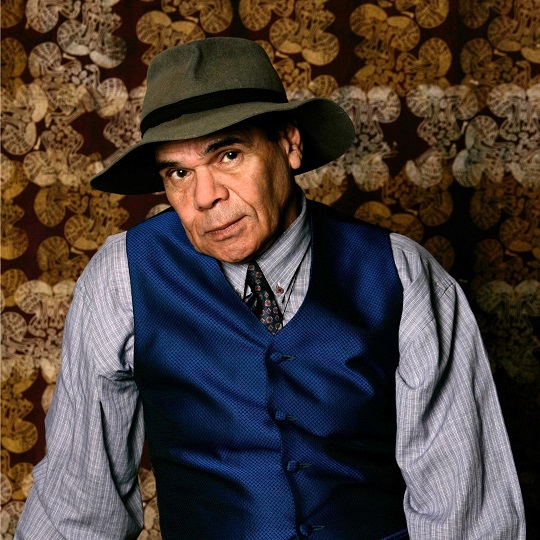 Portrait photo of Jeffrey Samuels by Sharon Hickey, Jeffrey looks at the camera almost smiling and wears a felt green/grey hat with a black brim, he wears a light grey long sleeve shirt underneath a silk navy blue waistcoat and a dark blue tie with red diamond shapes